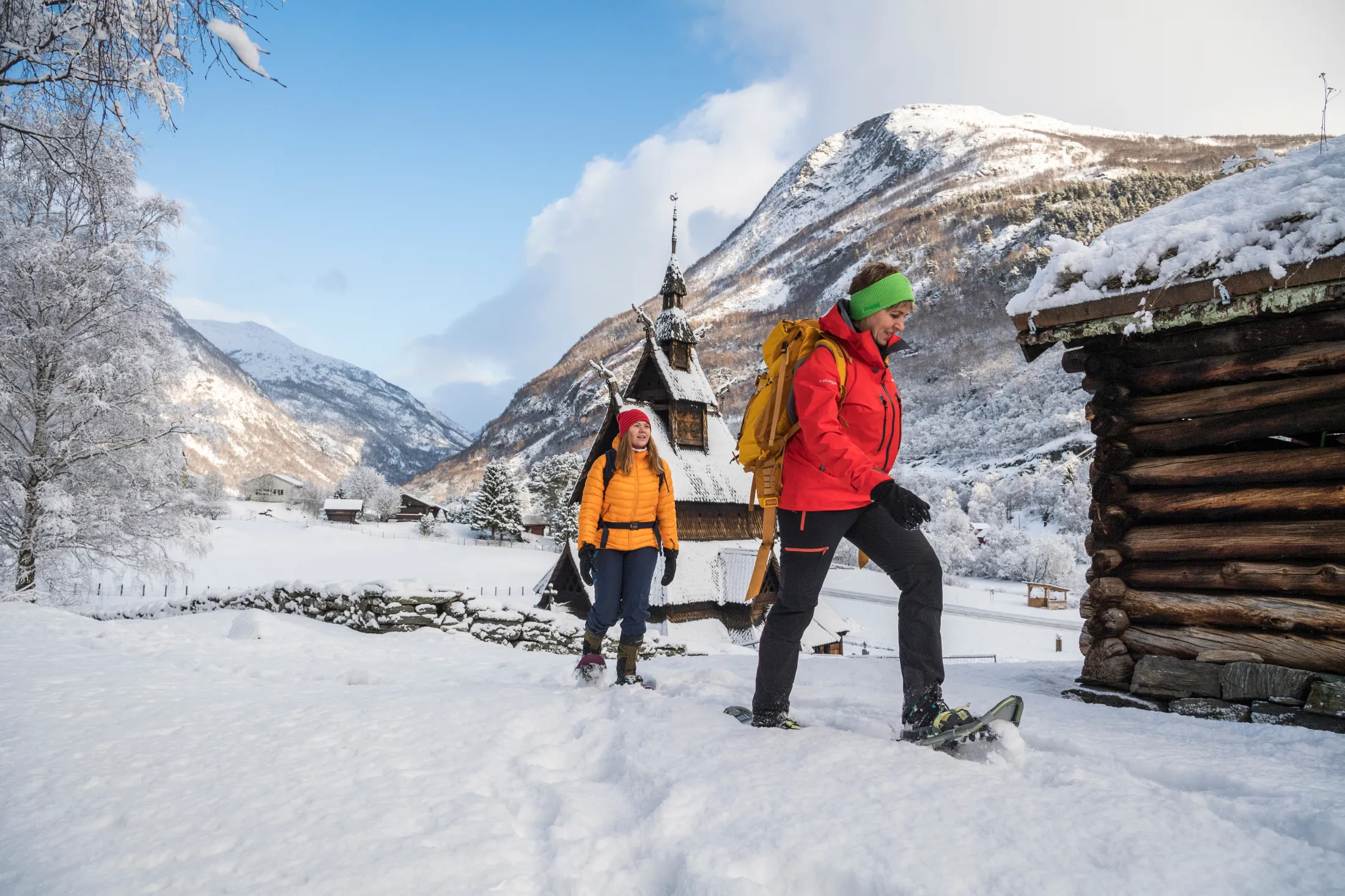 Two people snowshoeing in a snowy landscape with mountains and a traditional wooden church in the background.