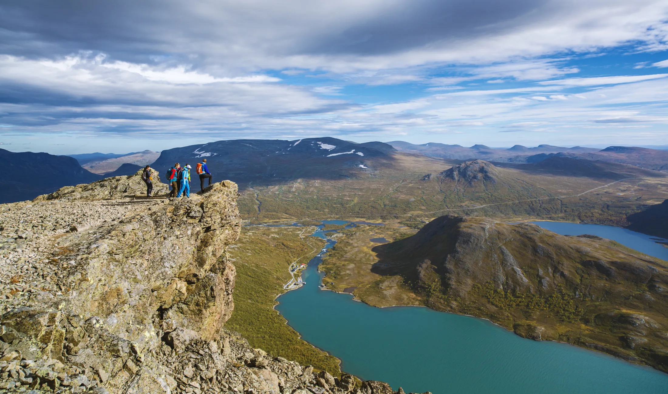 Hikers stand on a rocky mountain ledge overlooking a turquoise alpine lake and distant mountains under a cloudy sky.