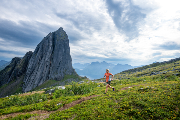 A trail runner runs through a lush green landscape beneath dramatic mountains under a cloudy sky.