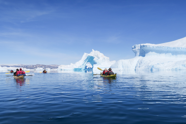 Several kayaks paddle on a clear, blue ocean toward a spectacular iceberg with a natural archway under a cloudless sky.