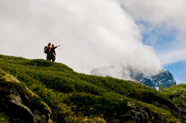 Two hikers stand on a grassy ridge, pointing at a mountain partially obscured by clouds.