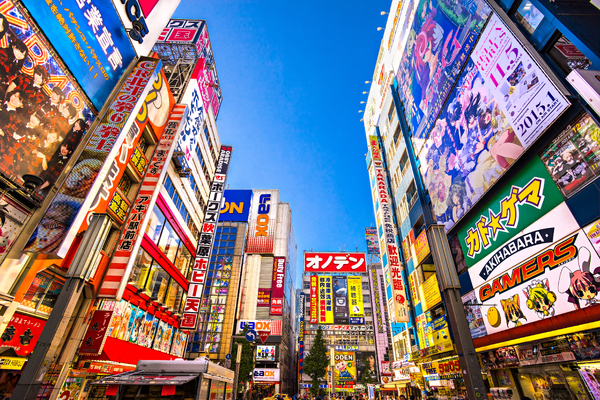 A low-angle shot looks up at towering buildings covered in colorful, bright anime billboards and neon signs in Tokyo's Akihabara district under a clear blue sky.