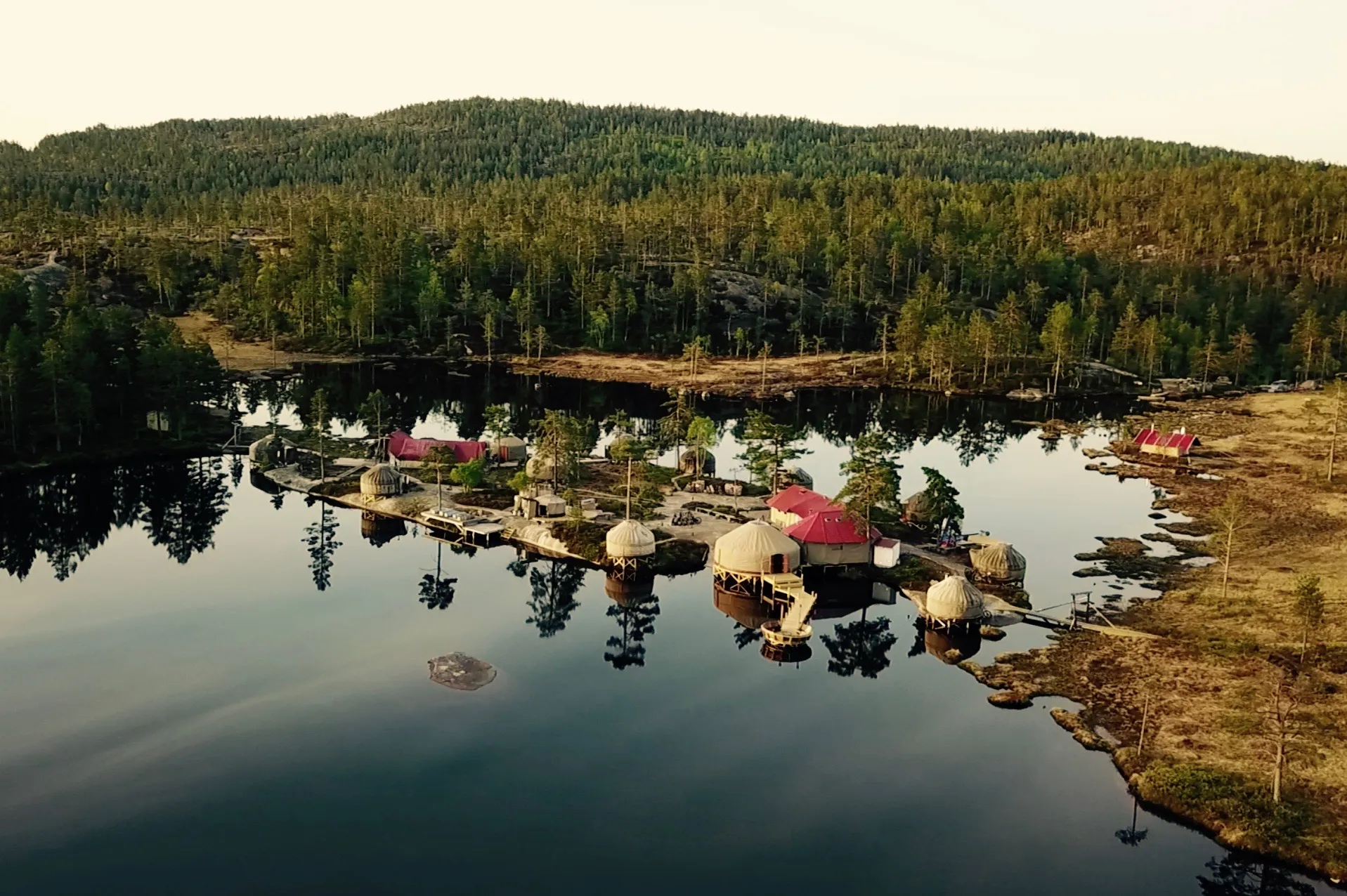 An aerial view of small domed structures and a larger red-roofed building on a narrow strip of land surrounded by a dark lake and green forest.