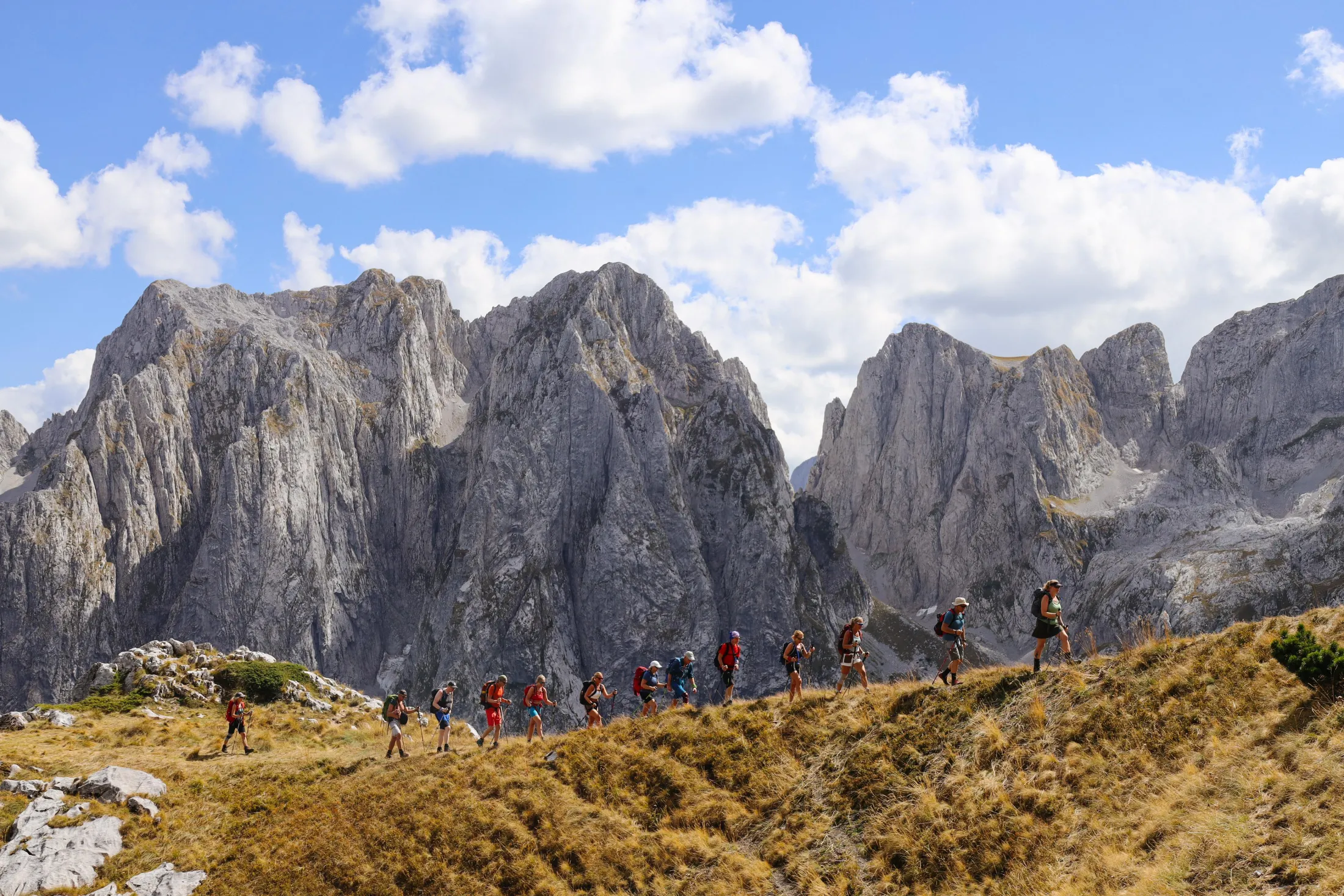 A line of hikers walking along a grassy mountain ridge with dramatic peaks and a blue sky in the background.