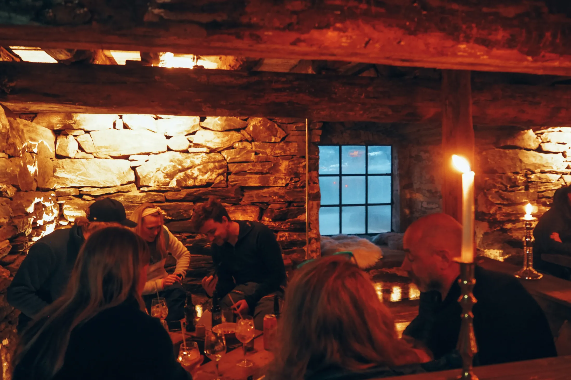 A group of people are gathered around a table inside a cozy, rustic cellar with stone walls and candlelight.
