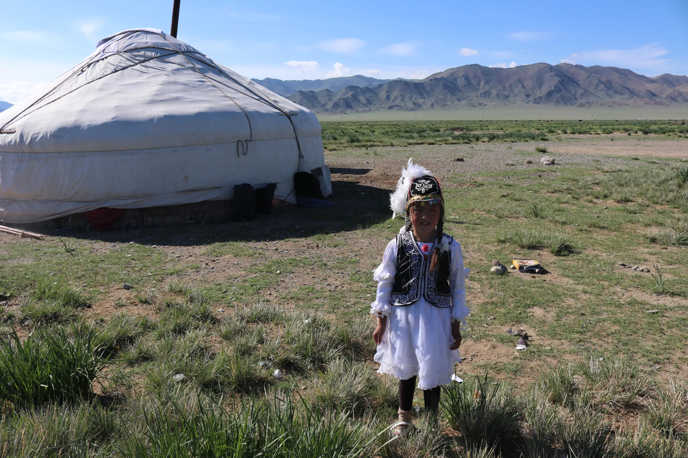A young girl in intricate traditional attire stands on a grassy field next to a large white yurt, with rolling mountains in the far distance under a bright blue sky.
