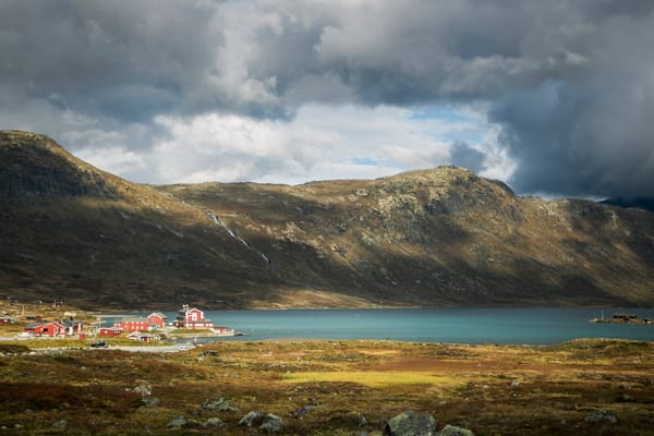 A mountain landscape with a lake, low vegetation, and a small settlement under a cloudy sky.