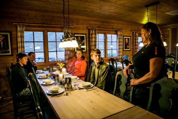 A group gathered around a wooden dining table inside a cabin, with a woman standing behind her chair.