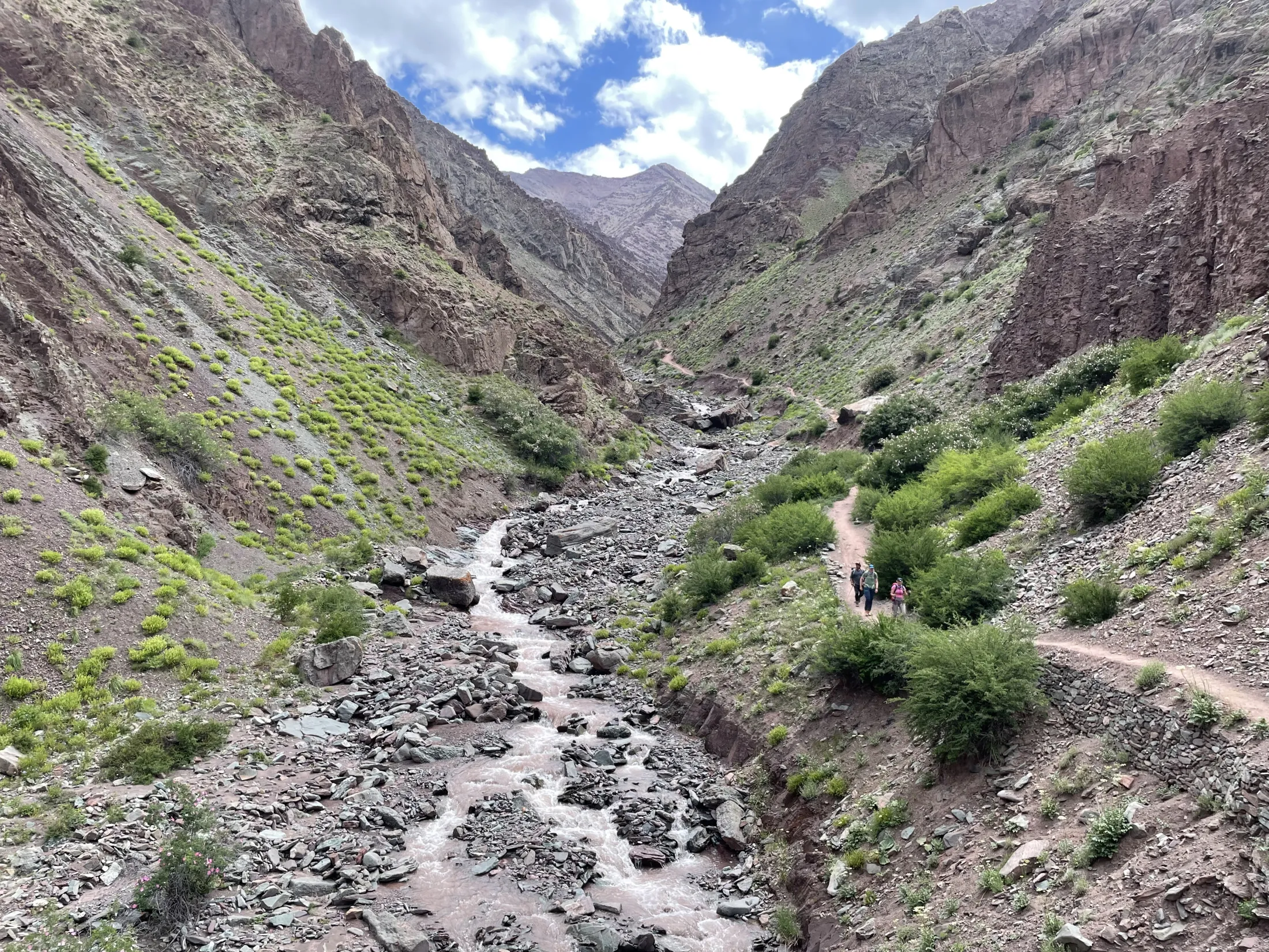 A few hikers walk along a narrow dirt path running parallel to a rocky, flowing river at the bottom of a steep, rugged mountain canyon.