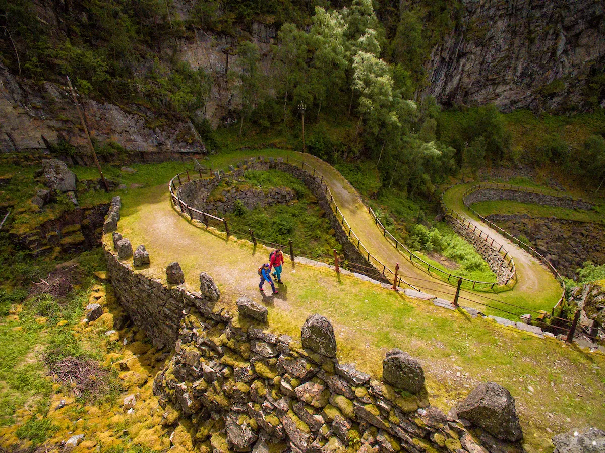 Two people walking along a winding dirt path in a lush green valley surrounded by rock walls.