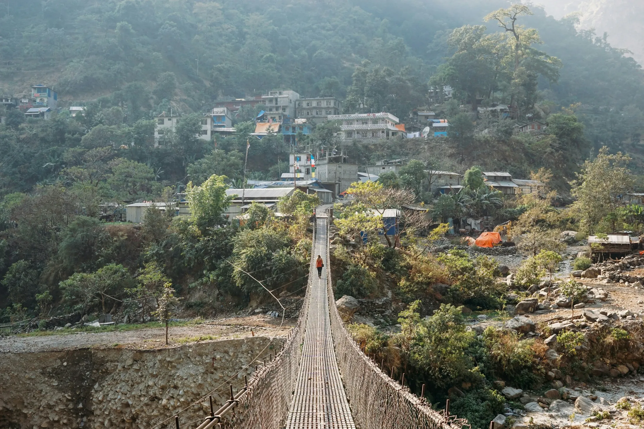 A person walks across a long suspension bridge leading toward a small hillside village nestled in a lush, mountainous landscape.