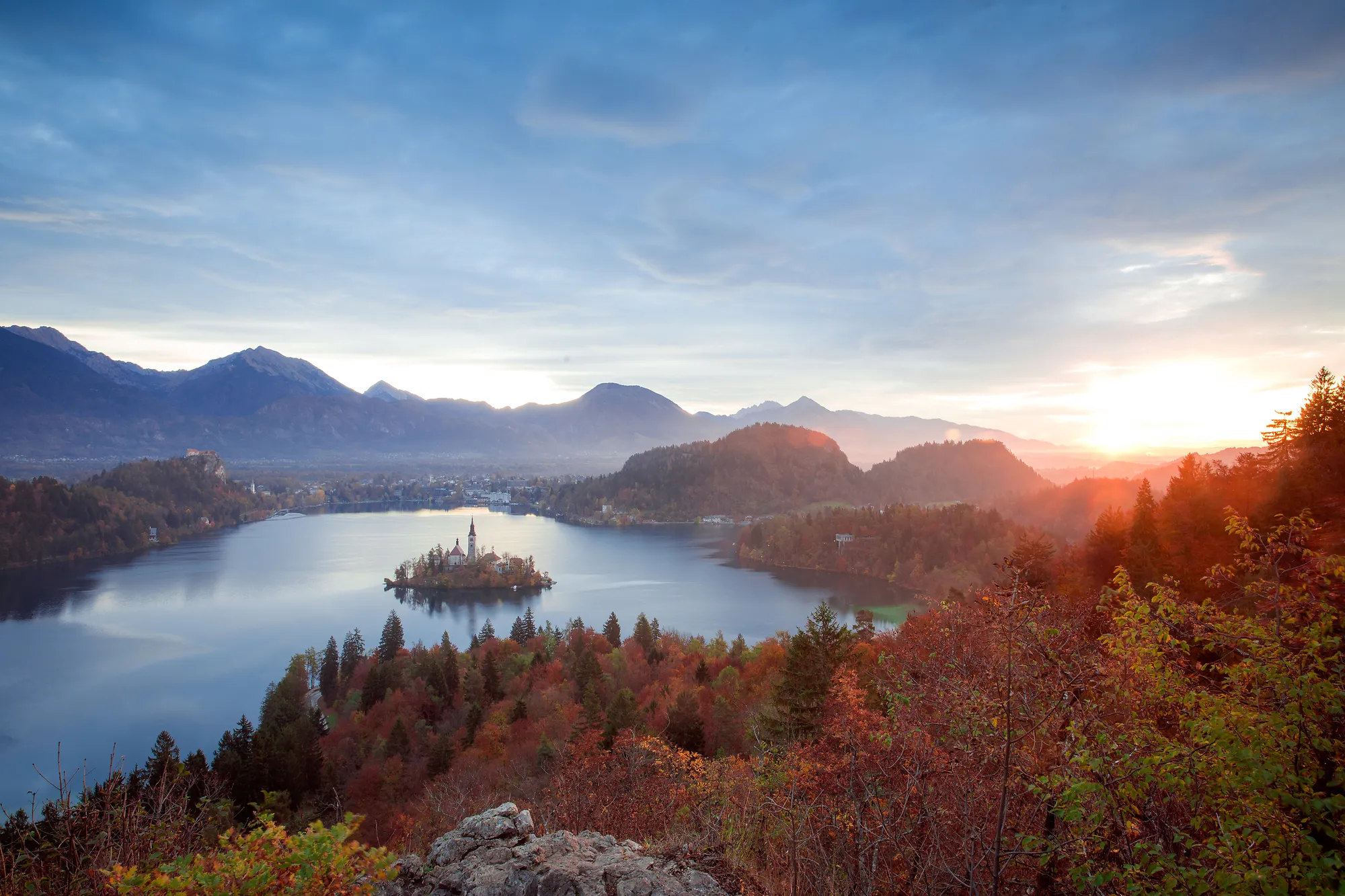 An aerial panoramic view looks over Lake Bled and its iconic island church during a soft sunrise, surrounded by autumn-colored forests and majestic mountains.