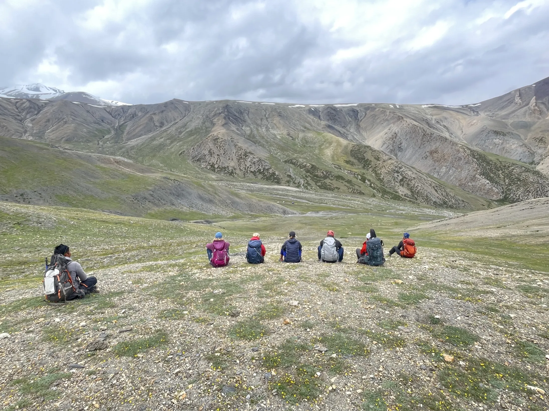 A group of hikers with backpacks sits on a grassy hill, resting and looking out over a vast mountain valley under a cloudy sky.
