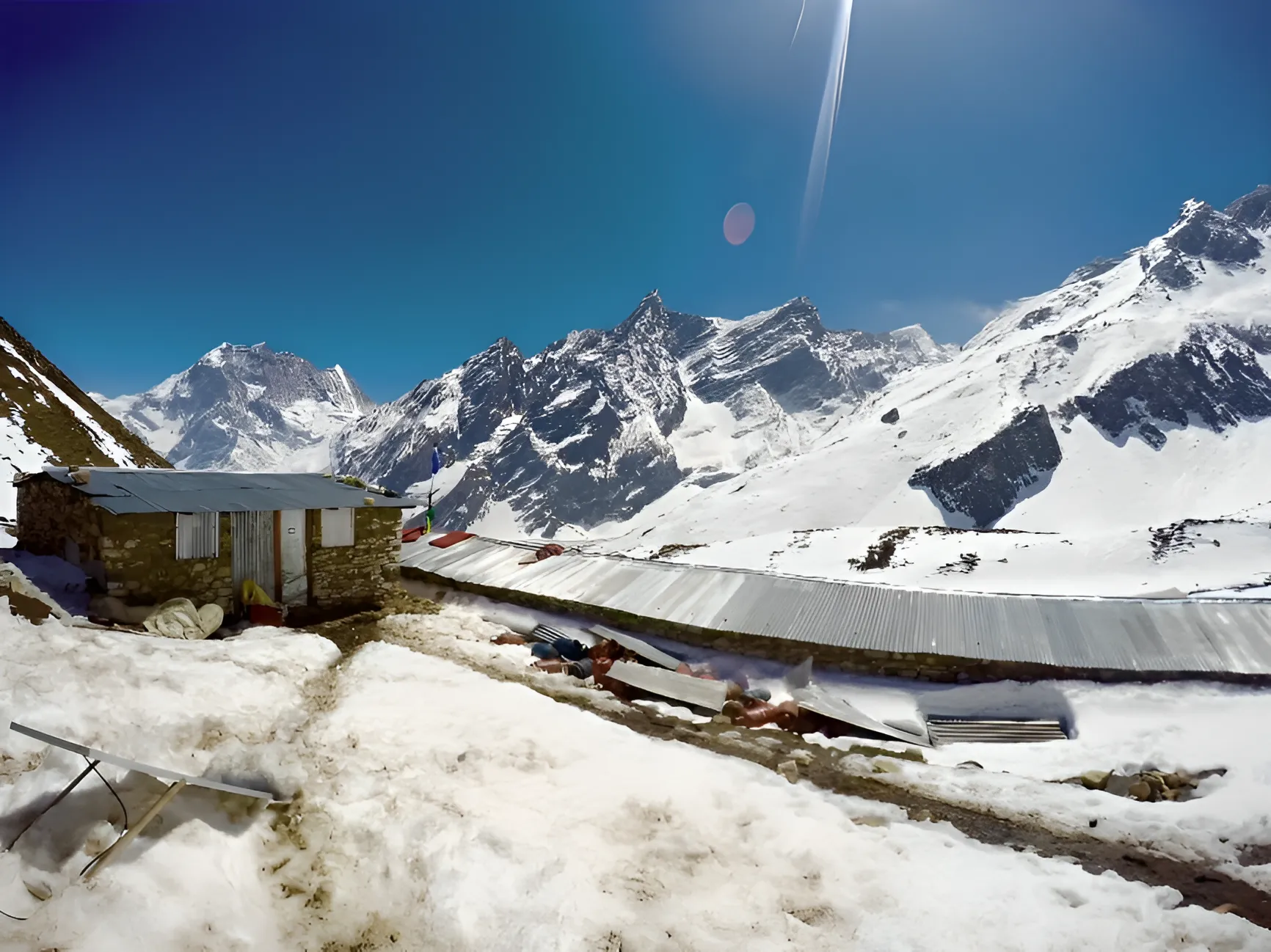Stone huts with corrugated metal roofs sit amidst deep snow, overlooking a dramatic landscape of jagged, snow-covered mountain peaks under a clear blue sky.