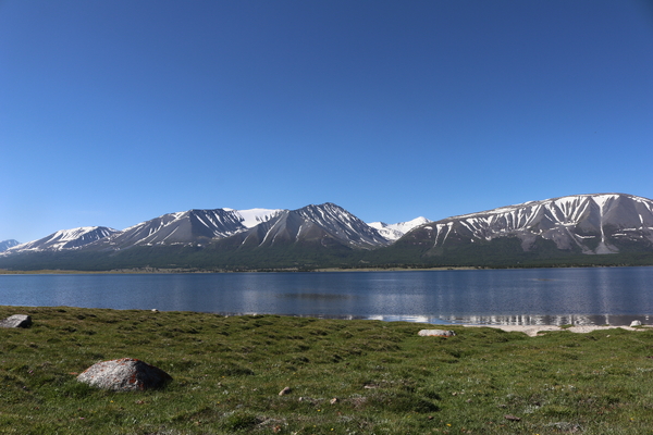 A calm, dark blue lake reflects snow-streaked mountains under a vast, cloudless blue sky, viewed from a grassy shore in the foreground.
