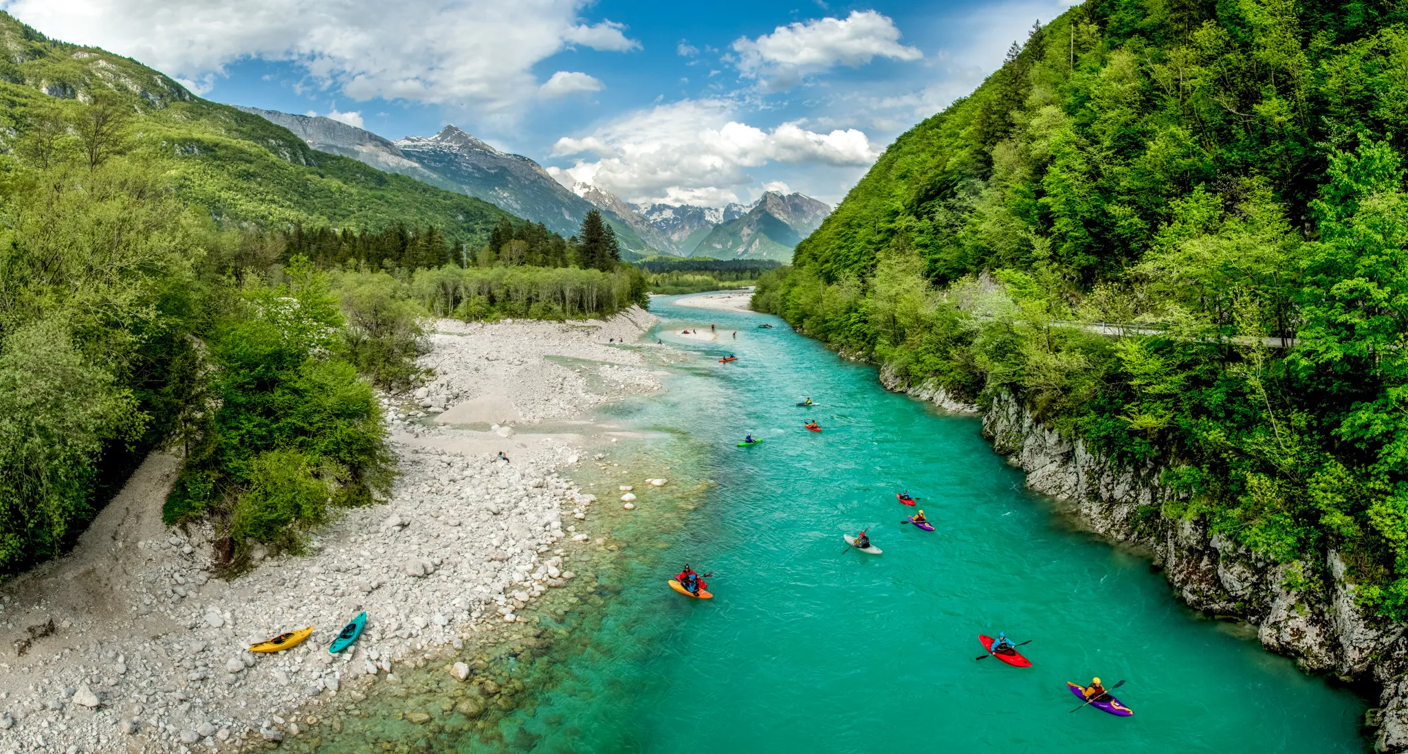 A vibrant turquoise river flows through a lush green valley, dotted with colorful kayakers and surrounded by steep, forested hills and distant, snow-capped mountains.