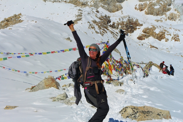 A smiling hiker with a backpack raises her arms in celebration on a snowy mountain pass decorated with colorful prayer flags.