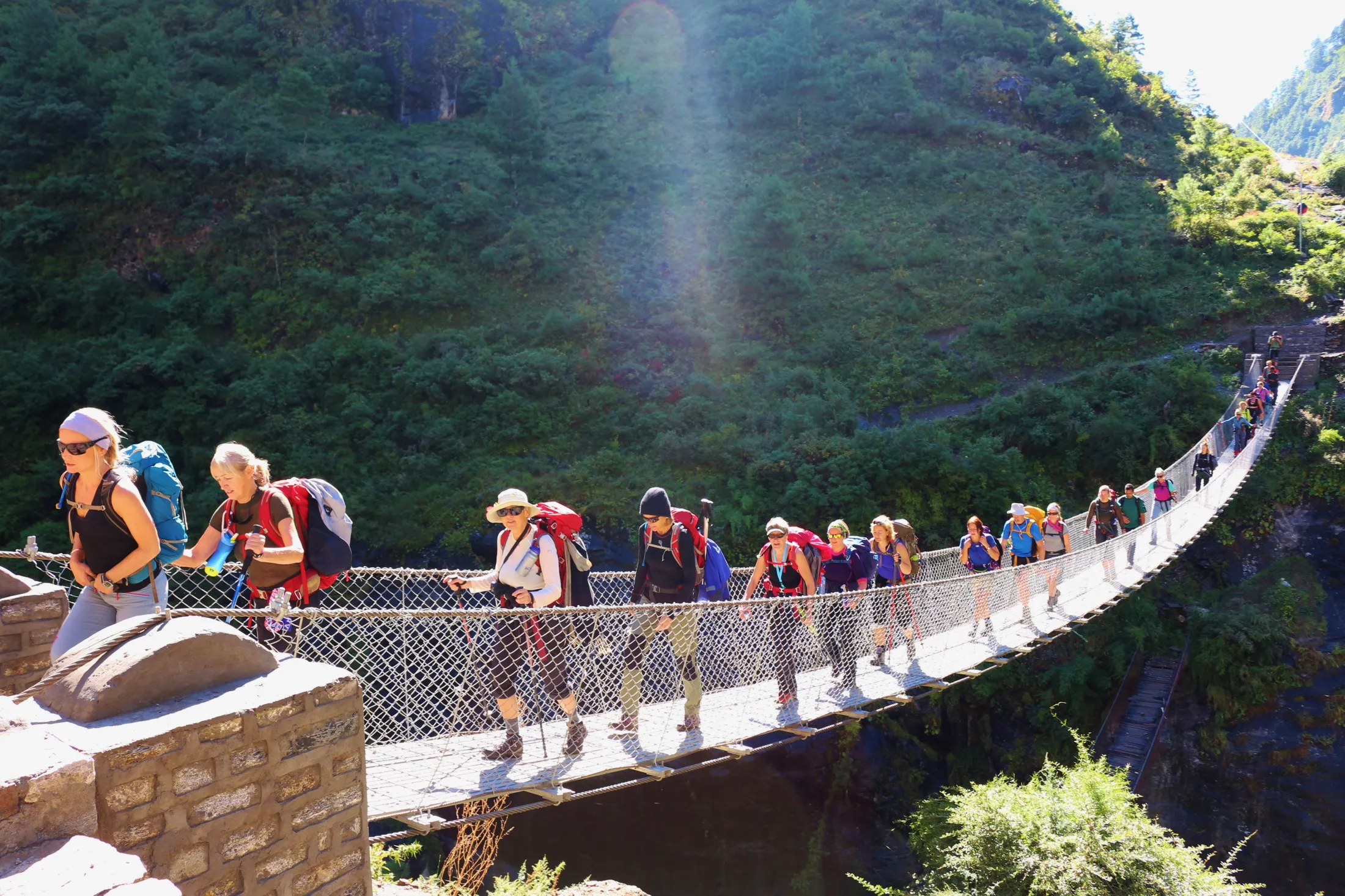 A long single-file line of hikers with backpacks crosses a suspension bridge over a gorge, with a lush green hillside in the background.