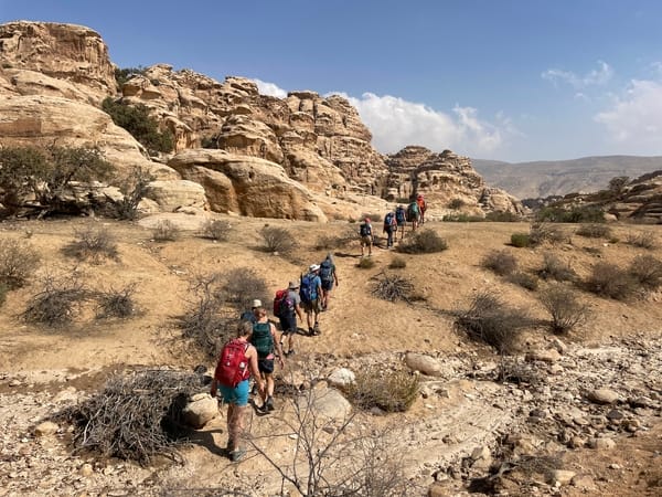 A group of hikers carrying backpacks walks in a line through a dry desert landscape toward towering sandstone cliffs.