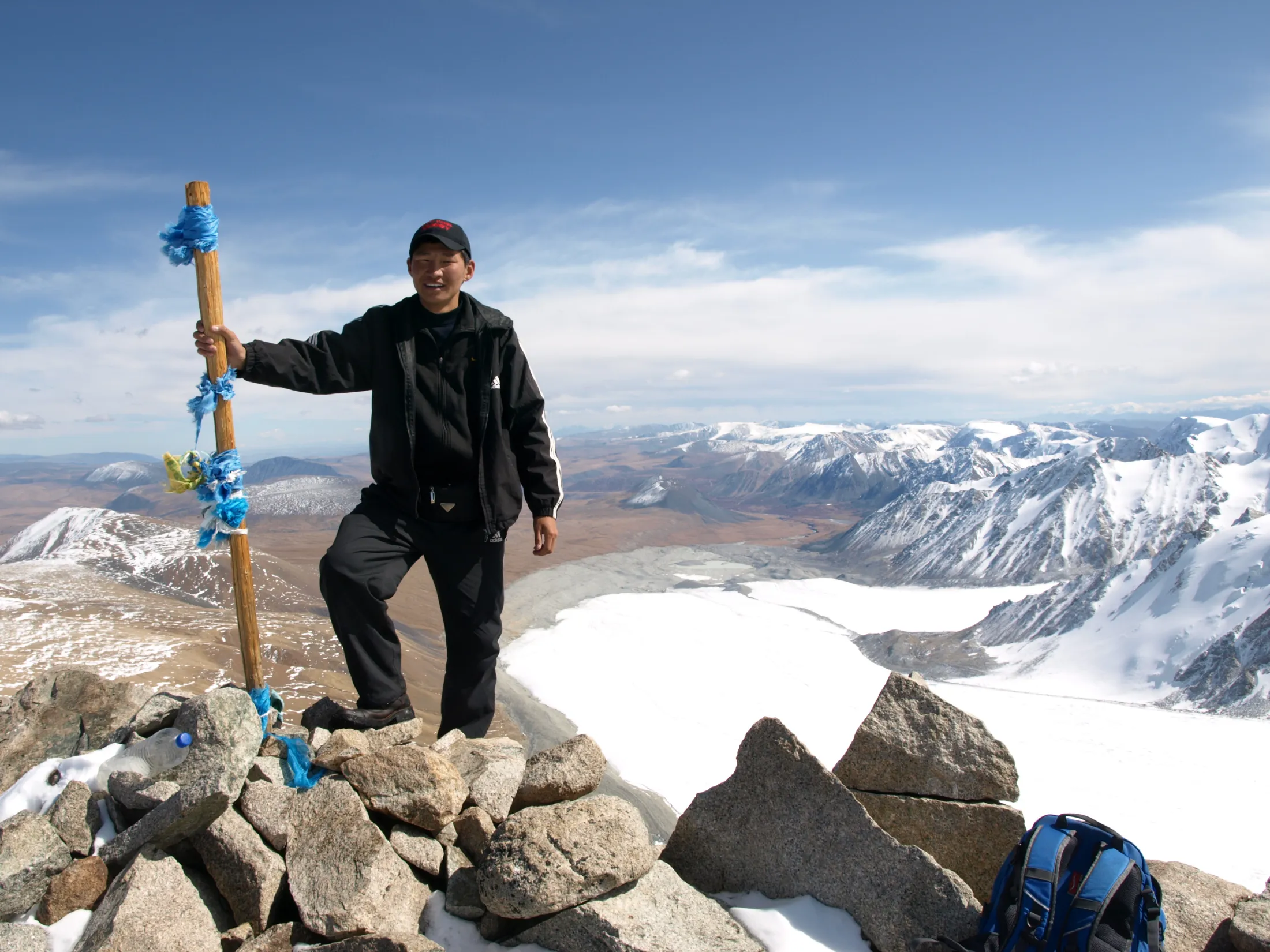 A man stands on a rocky mountain summit next to a wooden post decorated with blue ribbons, overlooking a vast glacier and snow-capped peaks under a blue sky.
