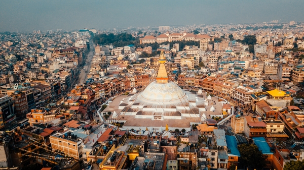 An aerial high-angle view shows the massive white dome and golden spire of the Boudhanath Stupa centered within a circular plaza, completely surrounded by the dense urban cityscape of Kathmandu under a hazy sky.