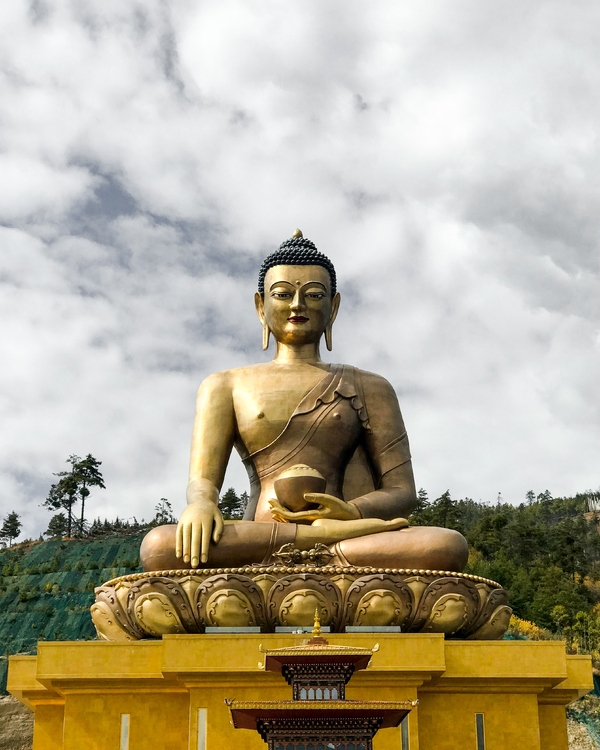 The giant golden Buddha Dordenma statue seated outdoors on a lotus throne against a cloudy sky and forested hills.