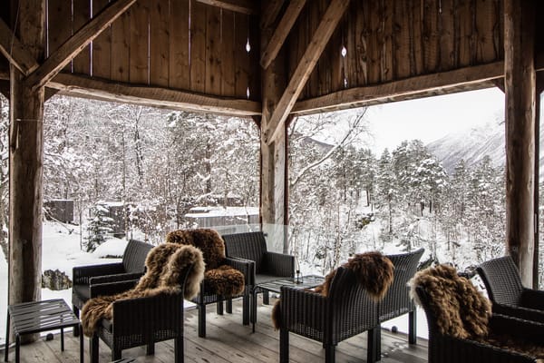 A cozy wooden porch with armchairs featuring fur throws, overlooking a snowy landscape.
