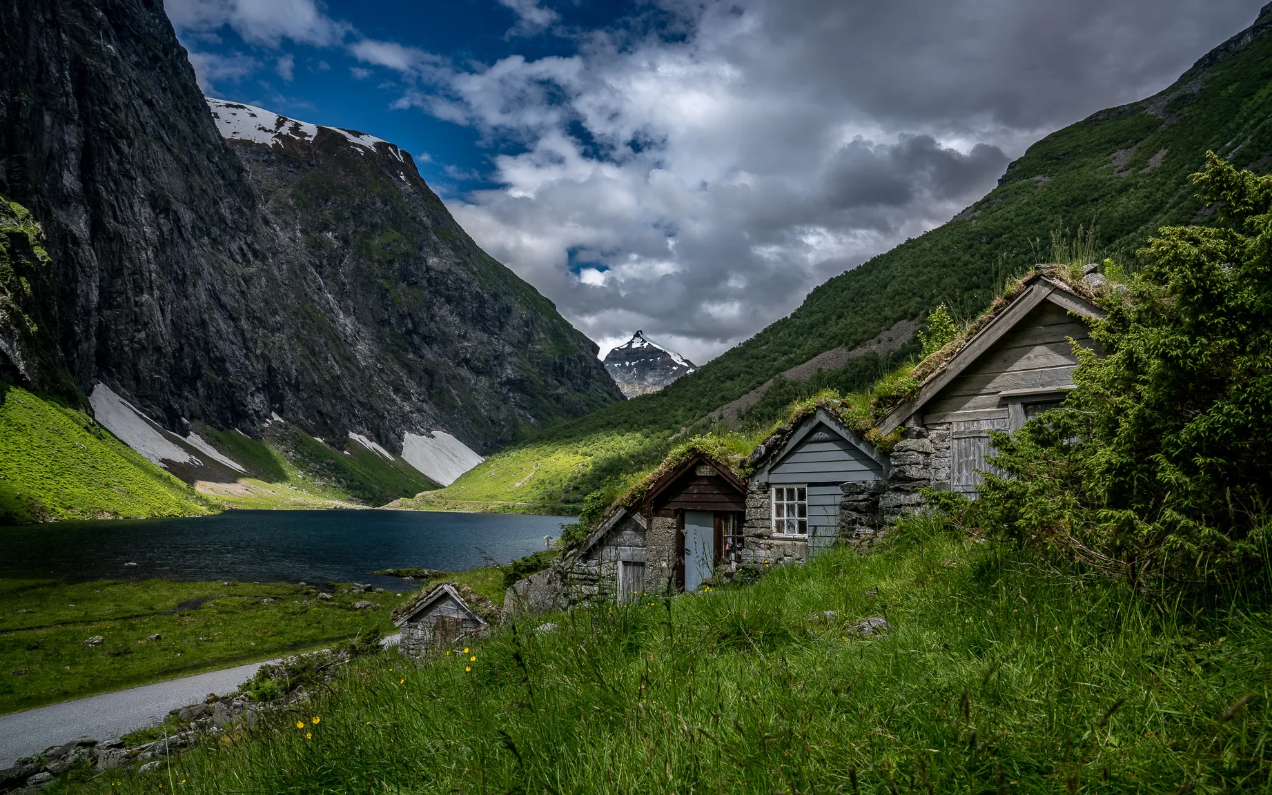 Traditional turf-roofed houses nestled in a lush, mountainous Norwegian valley beside a calm lake under a cloudy sky.