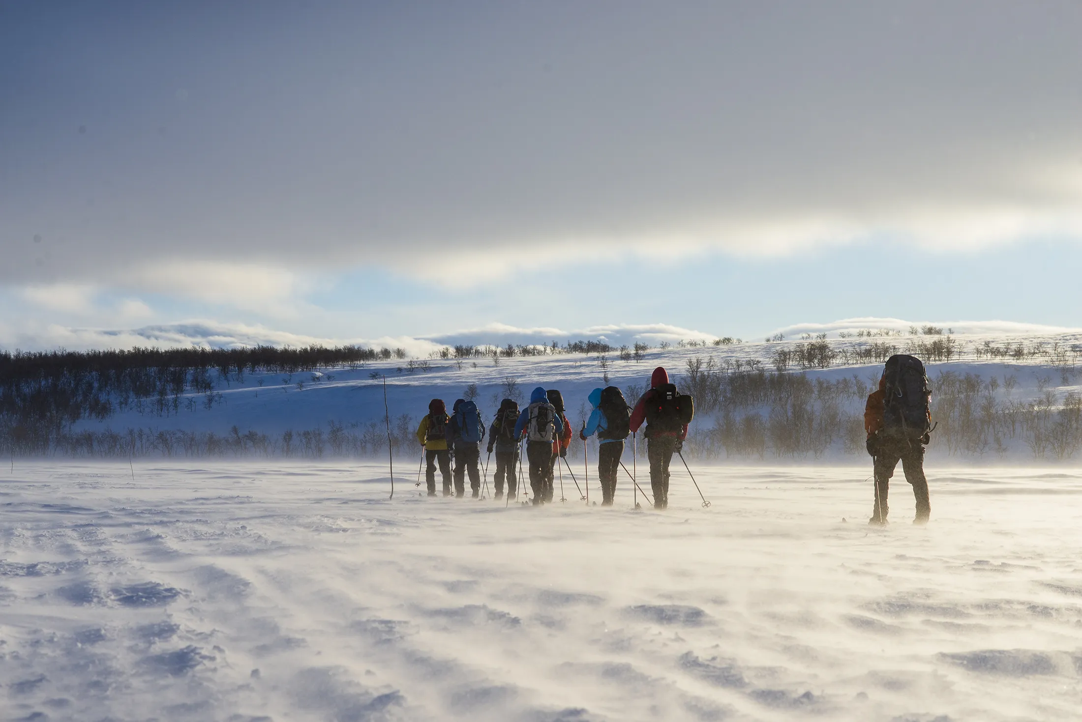 Cross-country skiers walking in a line across a snowy landscape under a cloudy sky.