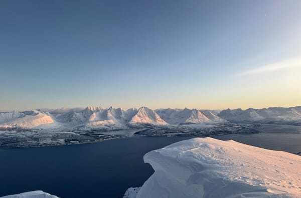 Aerial view of a snowy mountain ridge under a clear blue sky, overlooking a dark blue fjord with a coastal village below.