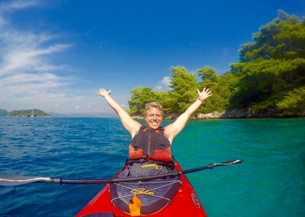 A smiling man wearing a life jacket sits in a red kayak on clear, turquoise sea water, celebrating with his arms raised high against a backdrop of a lush, pine-covered island under a bright blue sky.