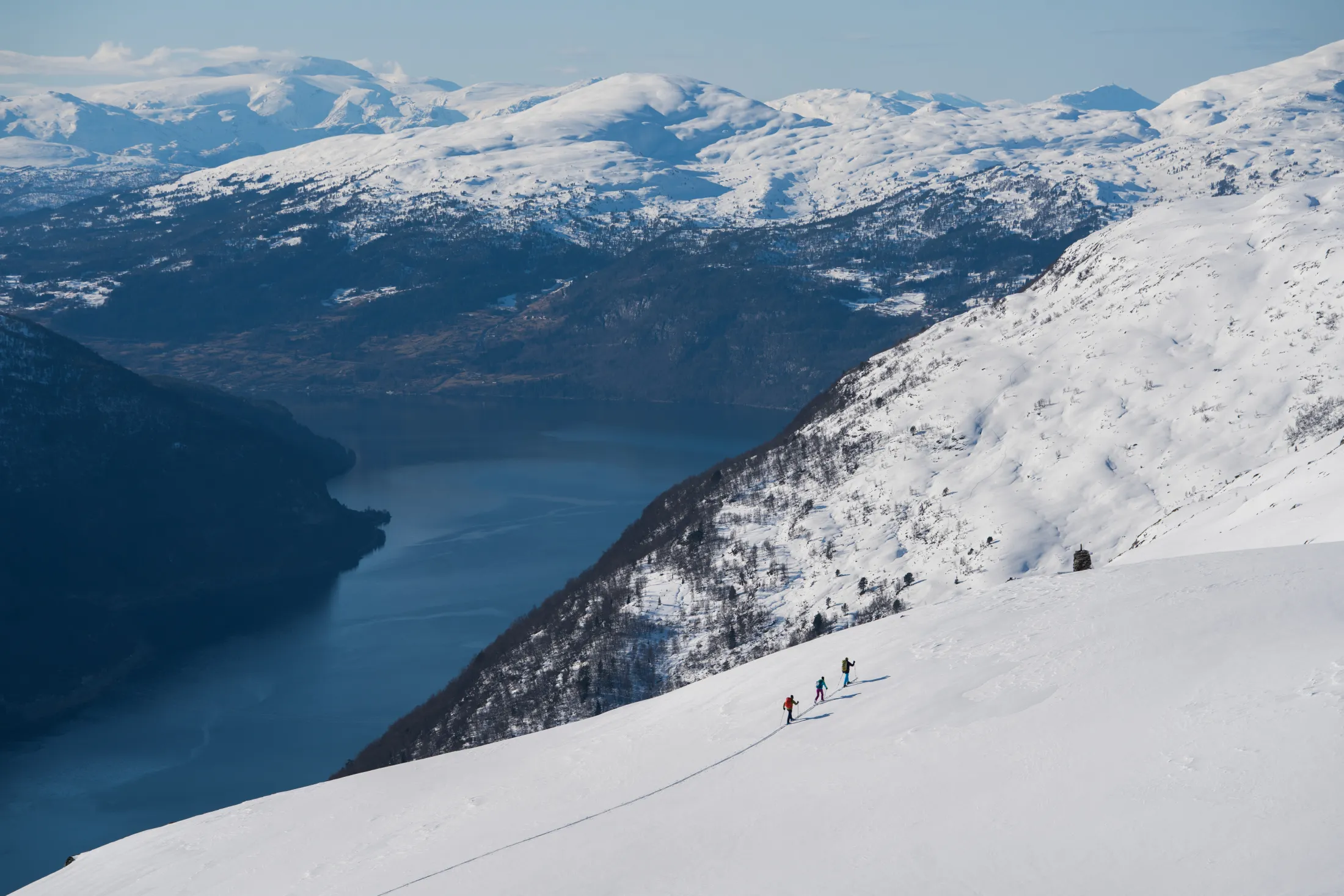 Three skiers climbing a snowy mountain slope overlooking a fjord and distant peaks.