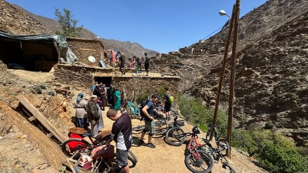 A group of cyclists resting with their bikes outside a rustic, stone-built mountain settlement under a clear blue sky.