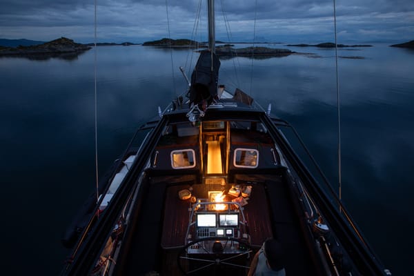 A sailboat navigates calm dark waters at dusk, islands in the background.