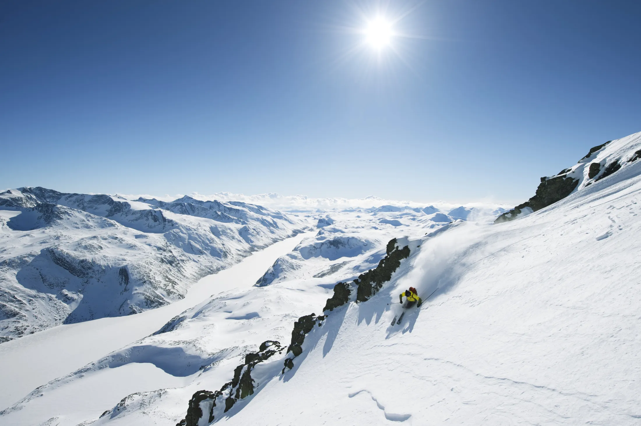 A skier in yellow jacket carving a turn down a steep, sunlit snowy mountain ridge with mountains in the background under a bright blue sky.