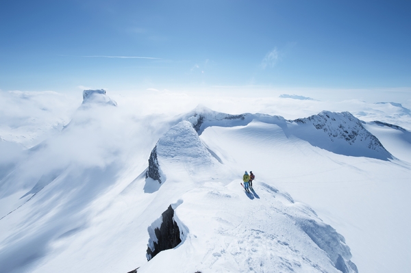 Aerial view of two skiers on a snowy mountain ridge under a bright blue sky.