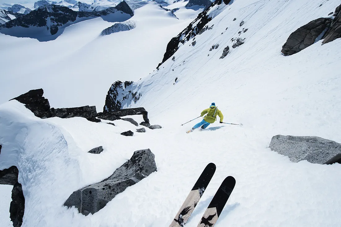 A skier in a bright yellow jacket navigates a steep, snow-covered mountain slope surrounded by rocks, with another set of skis visible in the foreground.