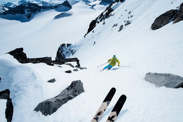 A skier in a bright yellow jacket navigates a steep, snow-covered mountain slope surrounded by rocks, with another set of skis visible in the foreground.