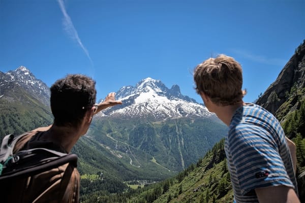 Two men look out over a deep valley as one points toward a massive, snow-covered mountain peak under a clear blue sky.