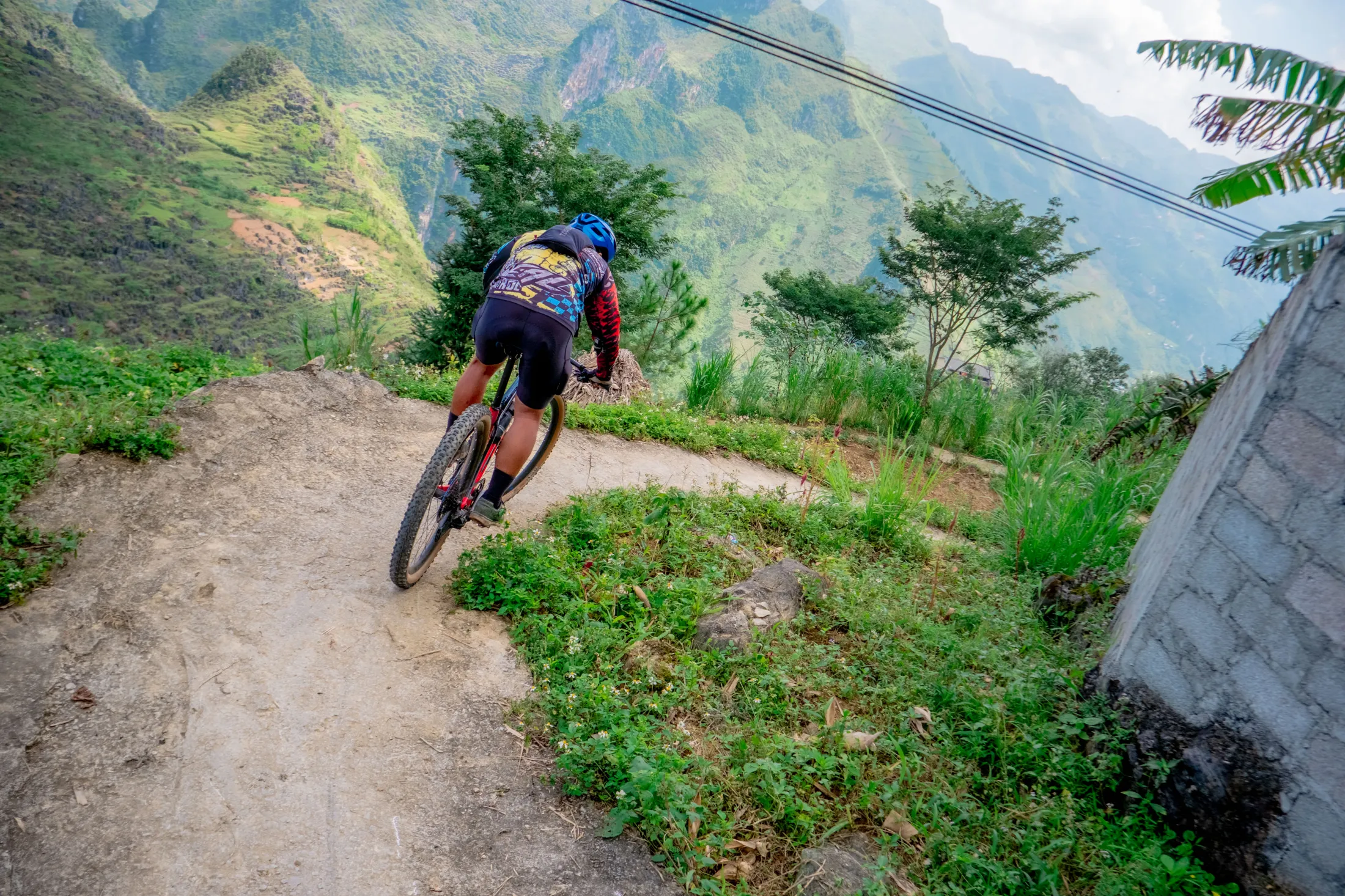A mountain biker cycling down a steep, winding dirt path in a lush, mountainous landscape.
