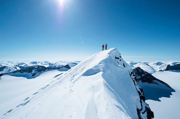 Two people standing on a snowy mountain peak under a bright blue sky.