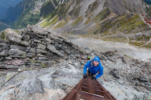 Looking down a steep, rocky mountainside, a smiling climber wearing a blue jacket and a blue helmet ascends a long metal ladder bolted to the cliff.