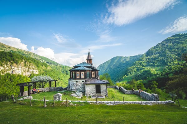 A historic stone and wooden church with a small steeple sits on a grassy hill, surrounded by a low stone wall and lush, green rolling mountains under a bright blue sky.