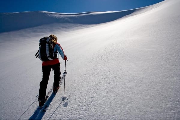 A skier with a backpack climbs a snow-covered mountain slope on a sunny day.
