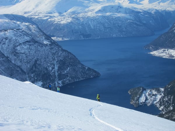 A skier descending a snowy mountain slope with a blue fjord and mountains in the background.