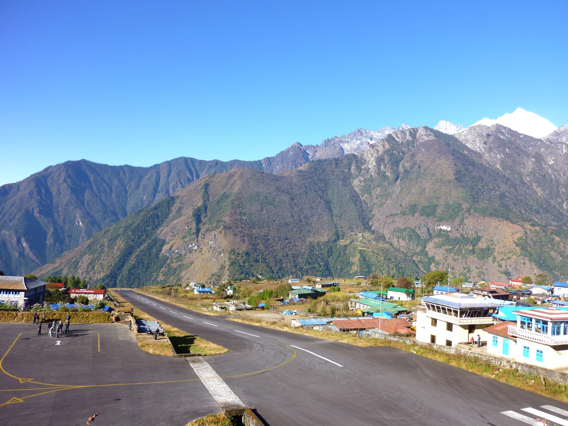 high-angle view looks down the short, sloped asphalt runway of Lukla Airport, surrounded by a mountain village and towering green hillsides under a clear blue sky.