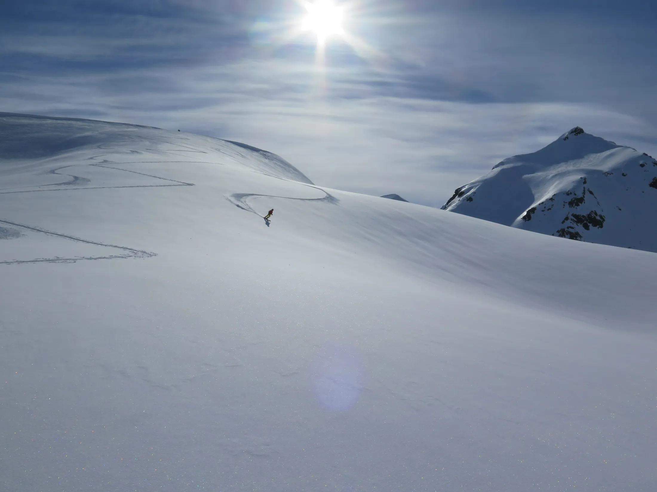 A lone skier on a vast snow-covered mountain slope under a bright sun.