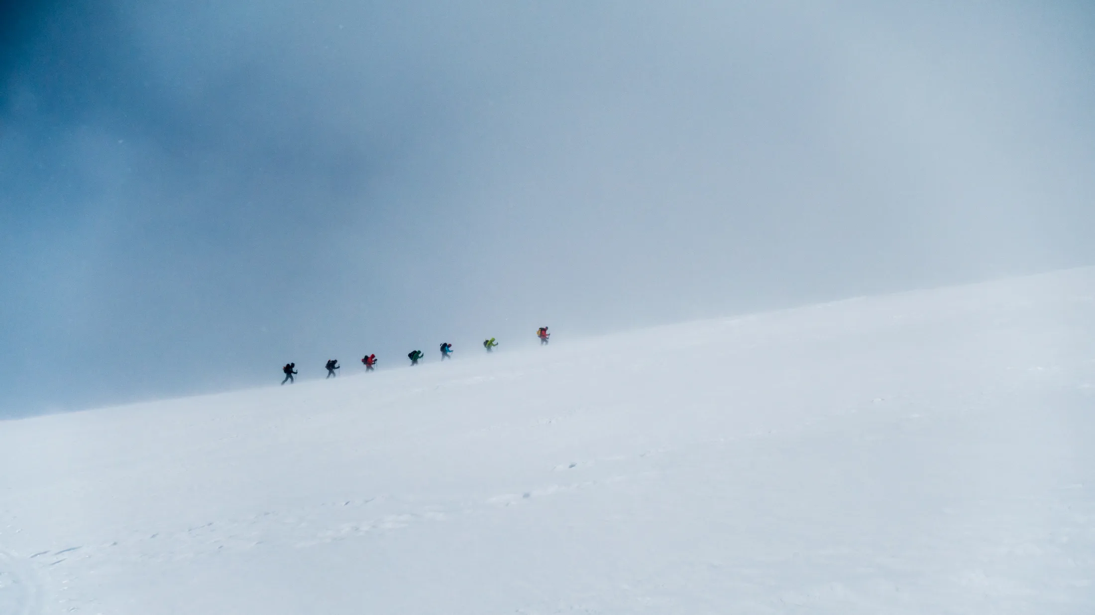 A line of small figures climbing a steep snow-covered mountain slope against a bright sky.