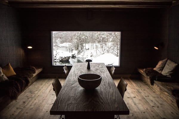 A cozy, dark-toned dining room with a long wooden table, built-in benches, and a large horizontal window overlooking a snowy landscape.