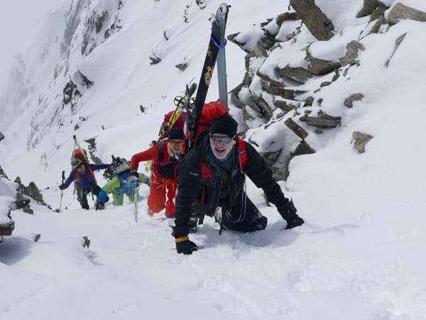 Four ski mountaineers crawl and climb up a steep, snowy slope with their skis attached to their backpacks alongside a rocky ridge.